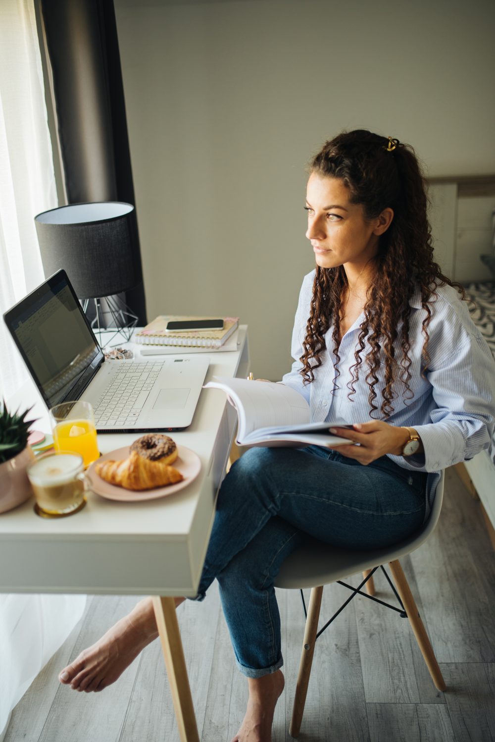 Young woman studying at a home desk with a laptop and breakfast nearby